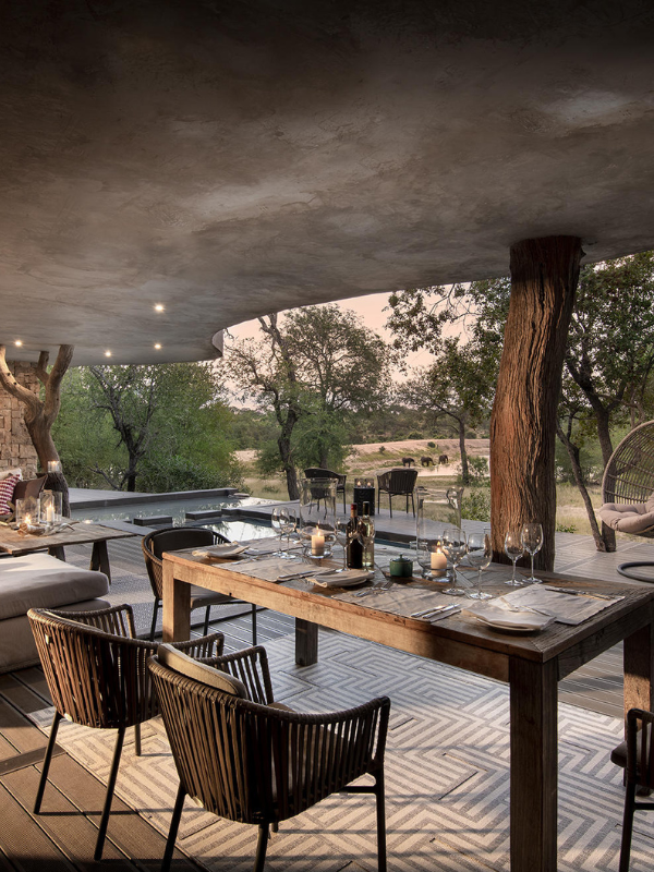 An outdoor dining deck under a modern, curved concrete overhang, featuring a long wooden table set for dinner with views of a waterhole where elephants are gathered in the distance.