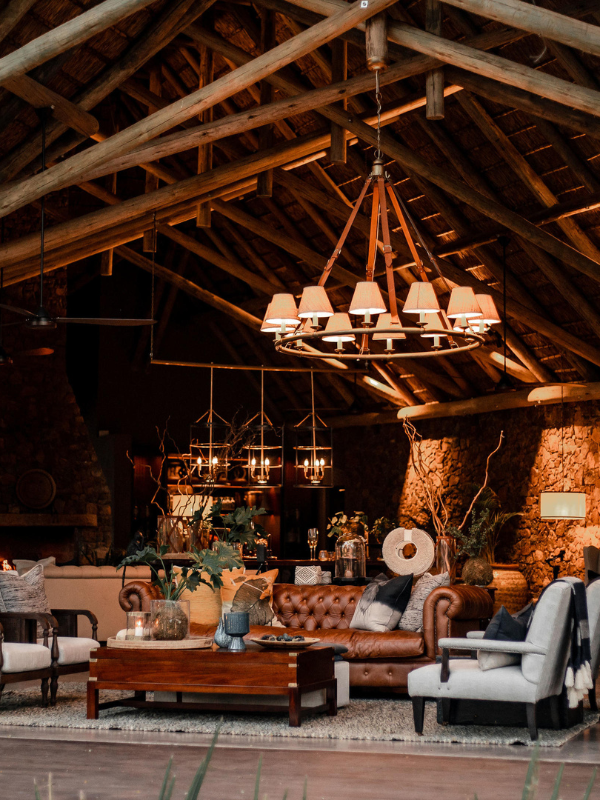 A sophisticated lounge area under a high thatched roof with exposed timber beams, centered around a leather Tufted sofa, a large circular chandelier, and a glowing stone fireplace.