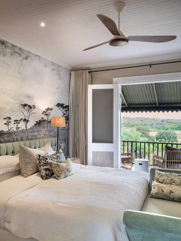 A classic safari bedroom with a vintage aesthetic, featuring a scenic tree-patterned mural behind the bed and white French doors that open onto a private veranda overlooking the bush.