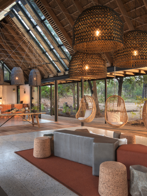 A bright, airy lodge interior with high vaulted ceilings, featuring large woven pendant lights, hanging wicker chairs, and modern minimalist furniture overlooking a sandy riverbed.