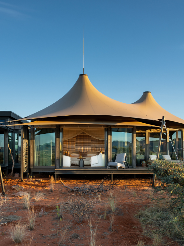 A contemporary safari tent with a unique, high-peaked tan canopy and glass walls, nestled in the red sand and sparse vegetation of the Kalahari
