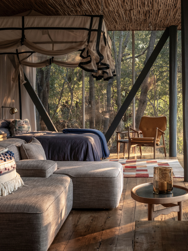 A luxury bedroom suite featuring a bed with a ruffled mosquito net and a plush gray sectional sofa, set against floor-to-ceiling glass walls that look directly into a dense green forest canopy.