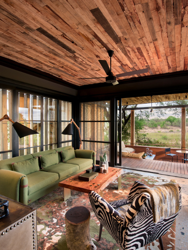 A stylish living area with a reclaimed wood ceiling and black-framed glass walls, featuring a green sofa, a live-edge wooden coffee table, and a zebra-print armchair.