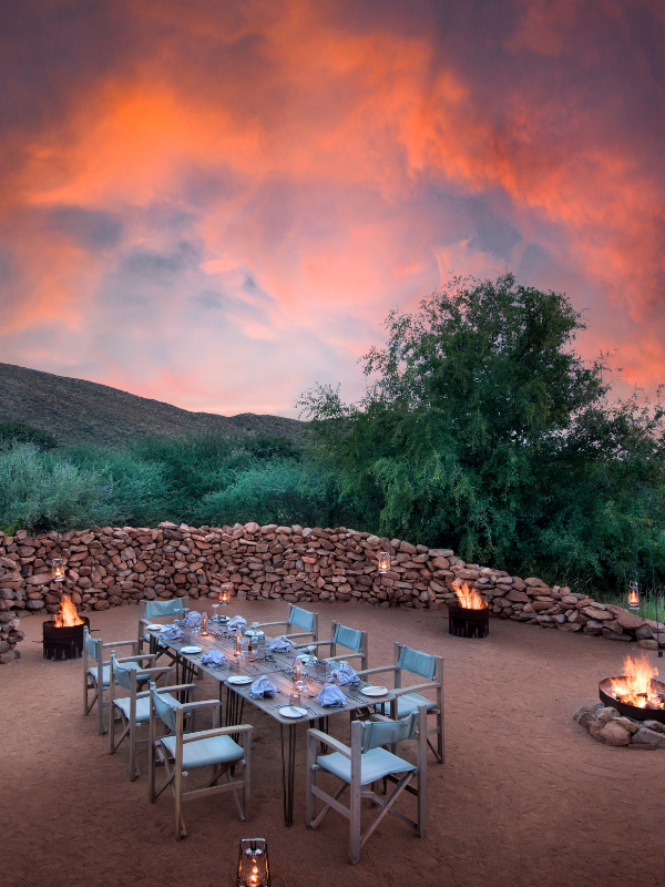 An intimate outdoor dining setup at dusk, featuring a long wooden table with white linens surrounded by safari chairs
